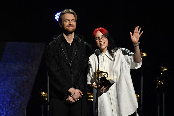 A man in black stands next to a woman in white at a microphone, accepting an award.