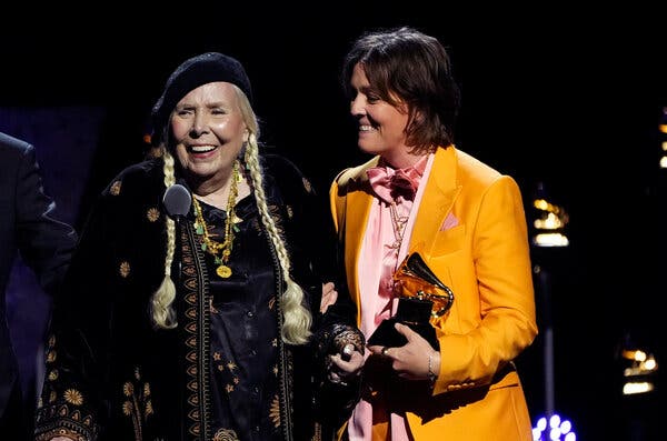 A woman in a beret with long blond braids stands at a microphone, beside a woman in a bright-yellow suit jacket.