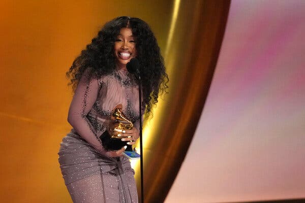 A woman in a sheer gray gown holds a Grammy in front of a microphone.